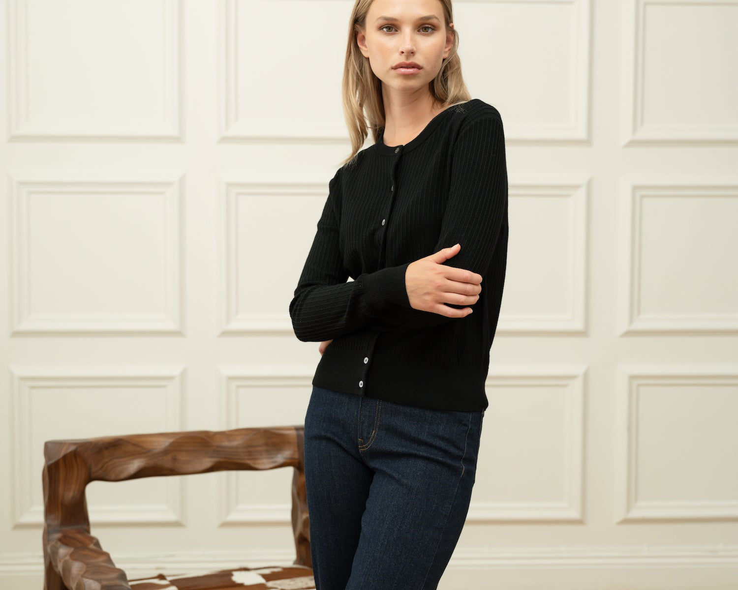 Woman wearing a Ninepine black merino wool cardigan with dark blue denim jeans against an off-white wainscoted wall