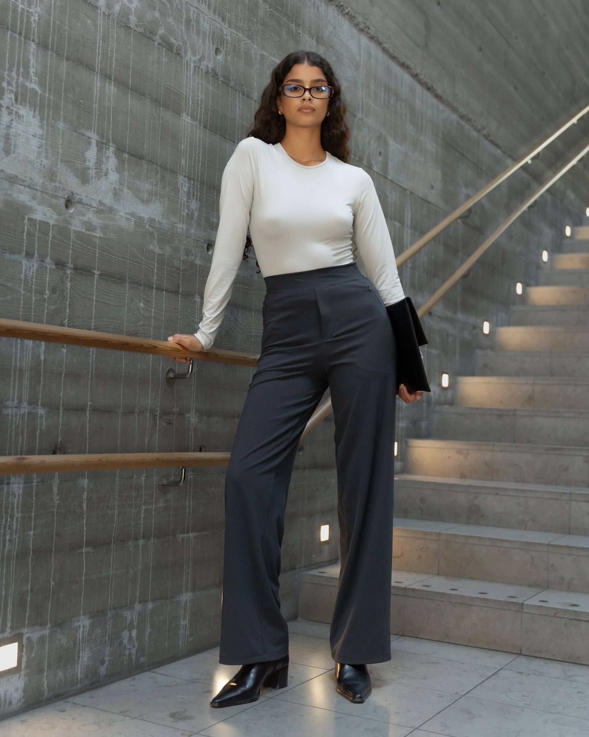 Woman walking up stairs wearing Ninepine Asana Relaxed trousers in dark grey styled with black boots for a city office look.